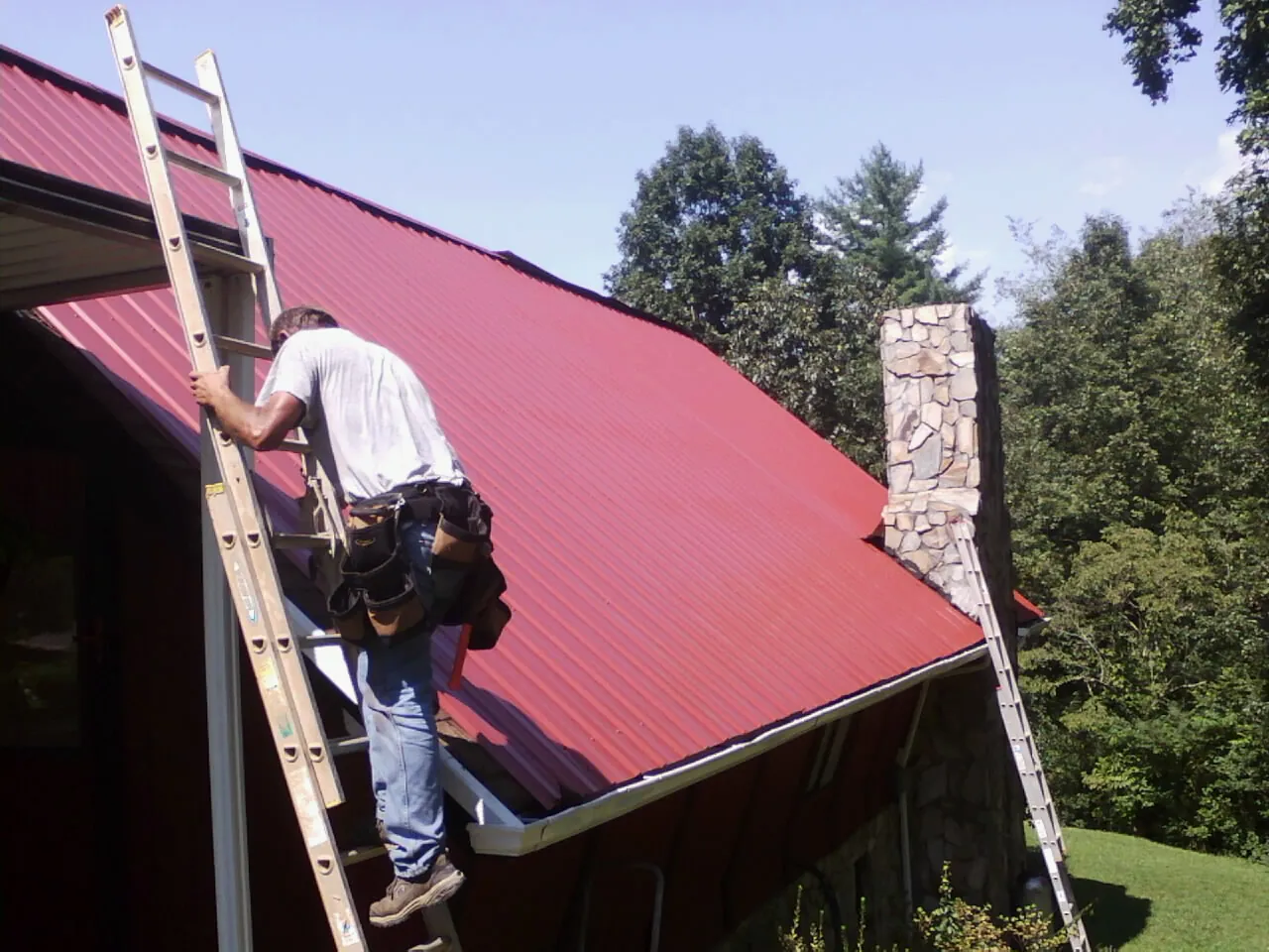 Roofer on a ladder beside a red metal roof and stone chimney — Soffit & Fascia Repair by Roofing Brothers in Kansas